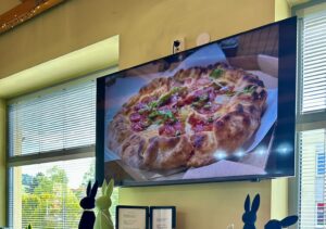 TV screen showing a pizza as a digital restaurant menu board above a piano in a cozy restaurant interior