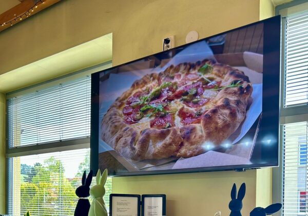 TV screen showing a pizza as a digital restaurant menu board above a piano in a cozy restaurant interior