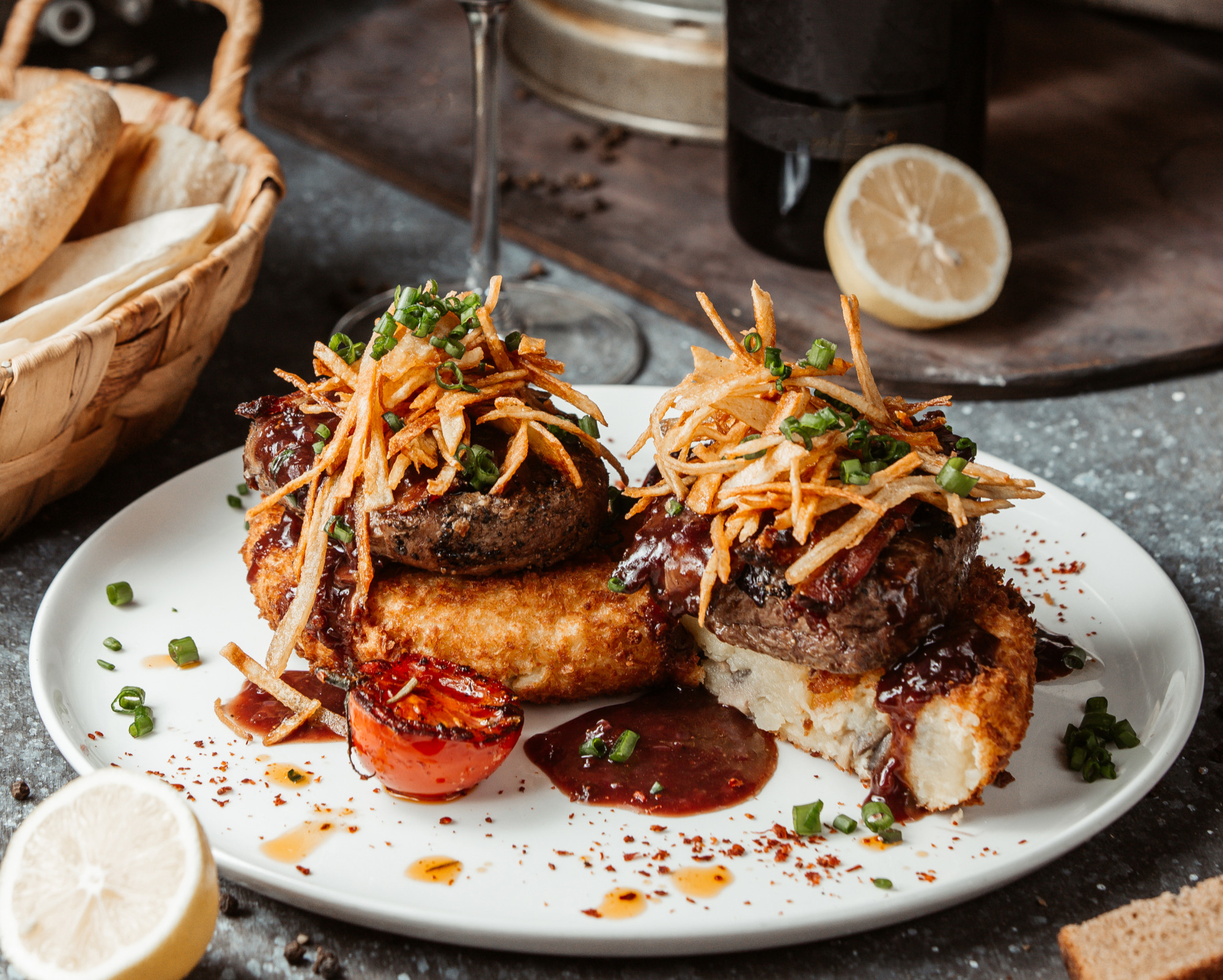A close-up of two gourmet beef patties topped with crispy fried onions and chives, served on golden-brown potato patties with a rich sauce and a grilled tomato on a white plate, with lemon slices and bread in the background.